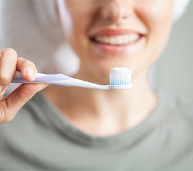 Woman in olive shirt blurry in background holding toothbrush with toothpaste on it in the foreground