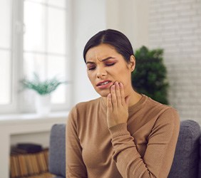 Woman in brown shirt in living room holding her hand to her sore teeth