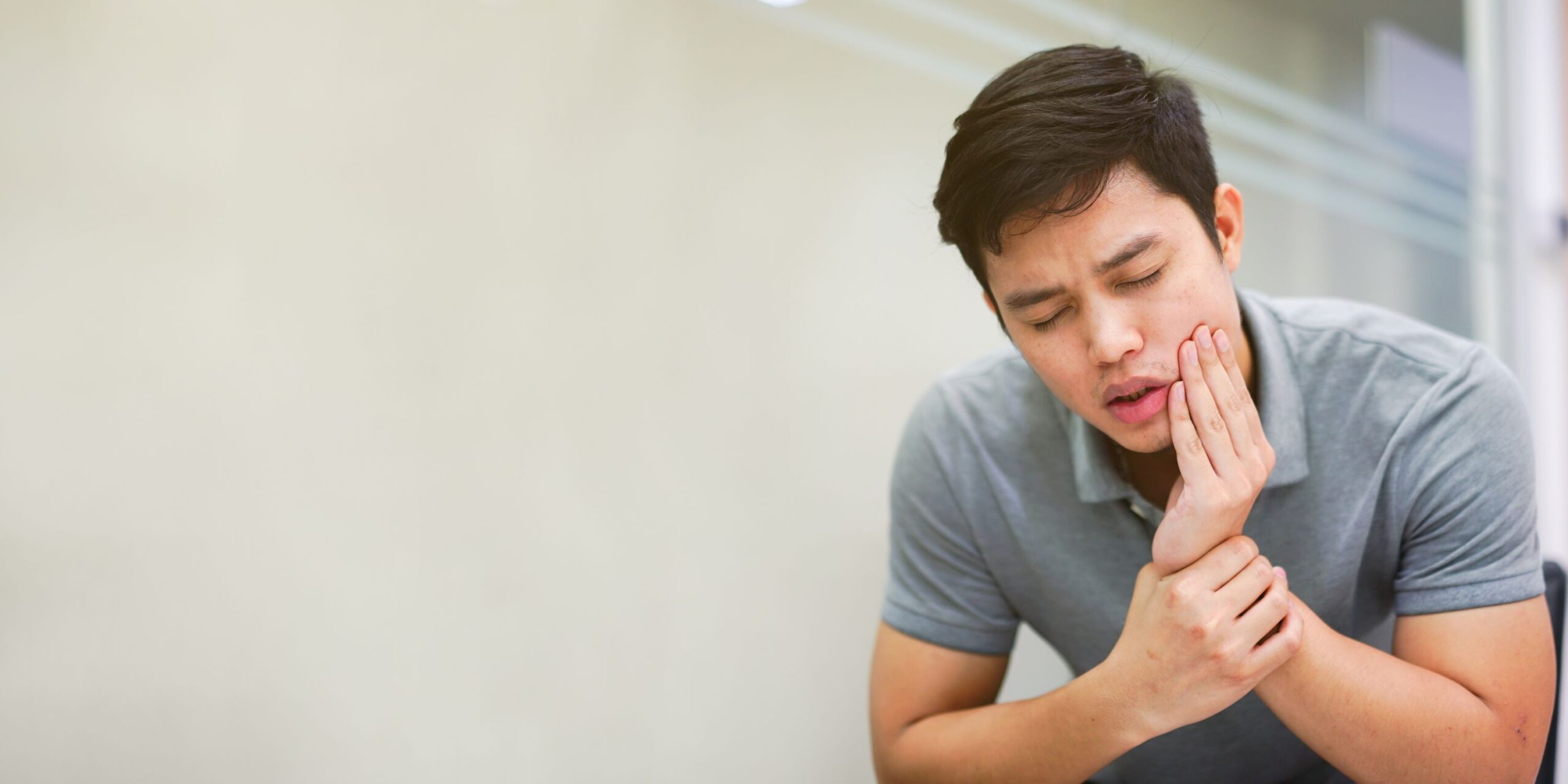 Man with brown hair in gray shirt with eyes closed holding hand to his jaw in pain